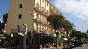 Um edifício de vários andares com varandas, um restaurante chamado 'B'LIVE' no andar térreo, mesas ao ar livre e céu claro.