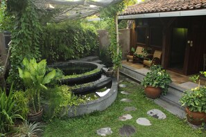 Un jardín con un estanque circular, plantas en macetas y un área de estar techada.