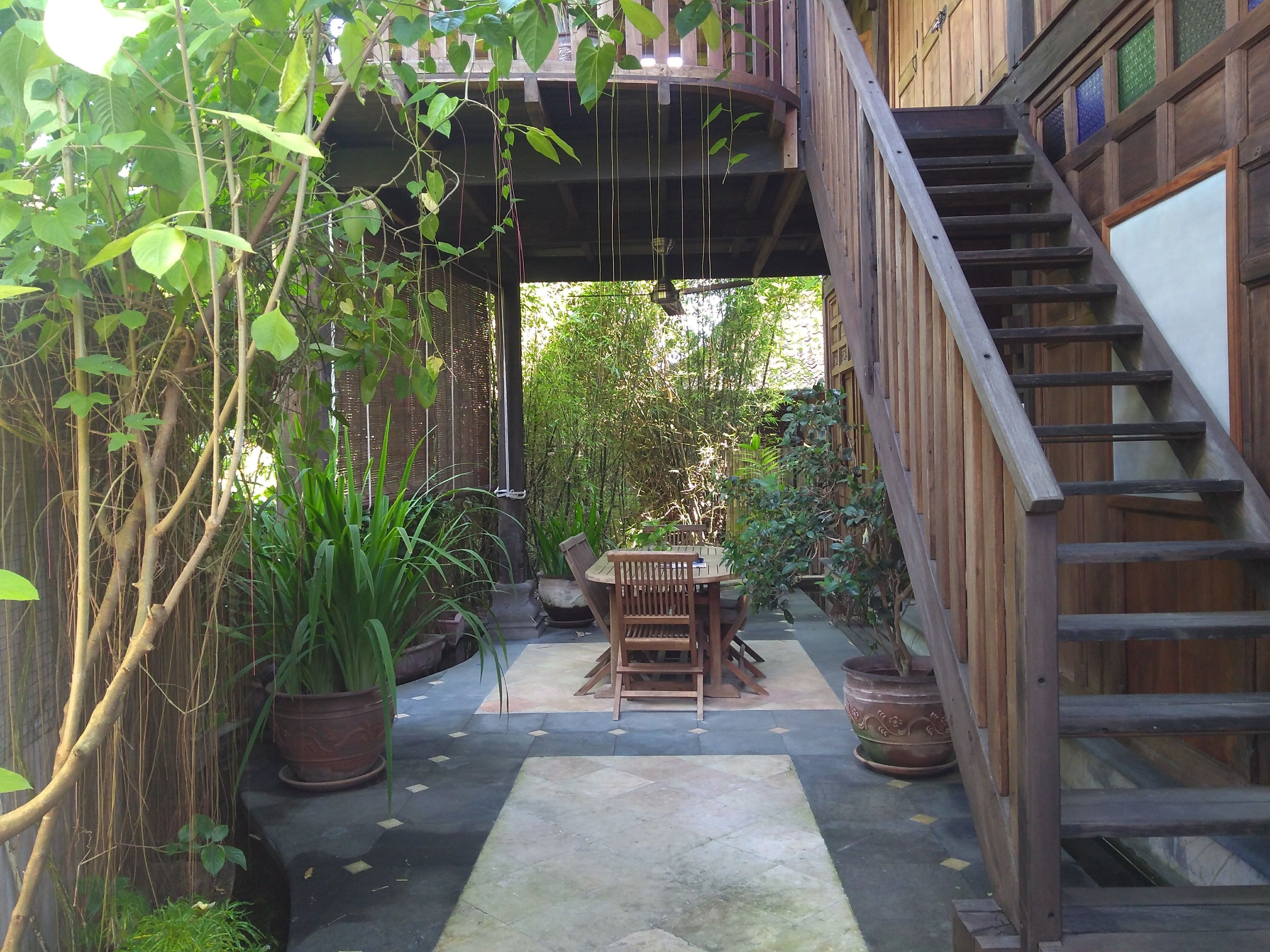 A wooden staircase leading to a covered outdoor area with a table and chairs, surrounded by potted plants and a bamboo fence.