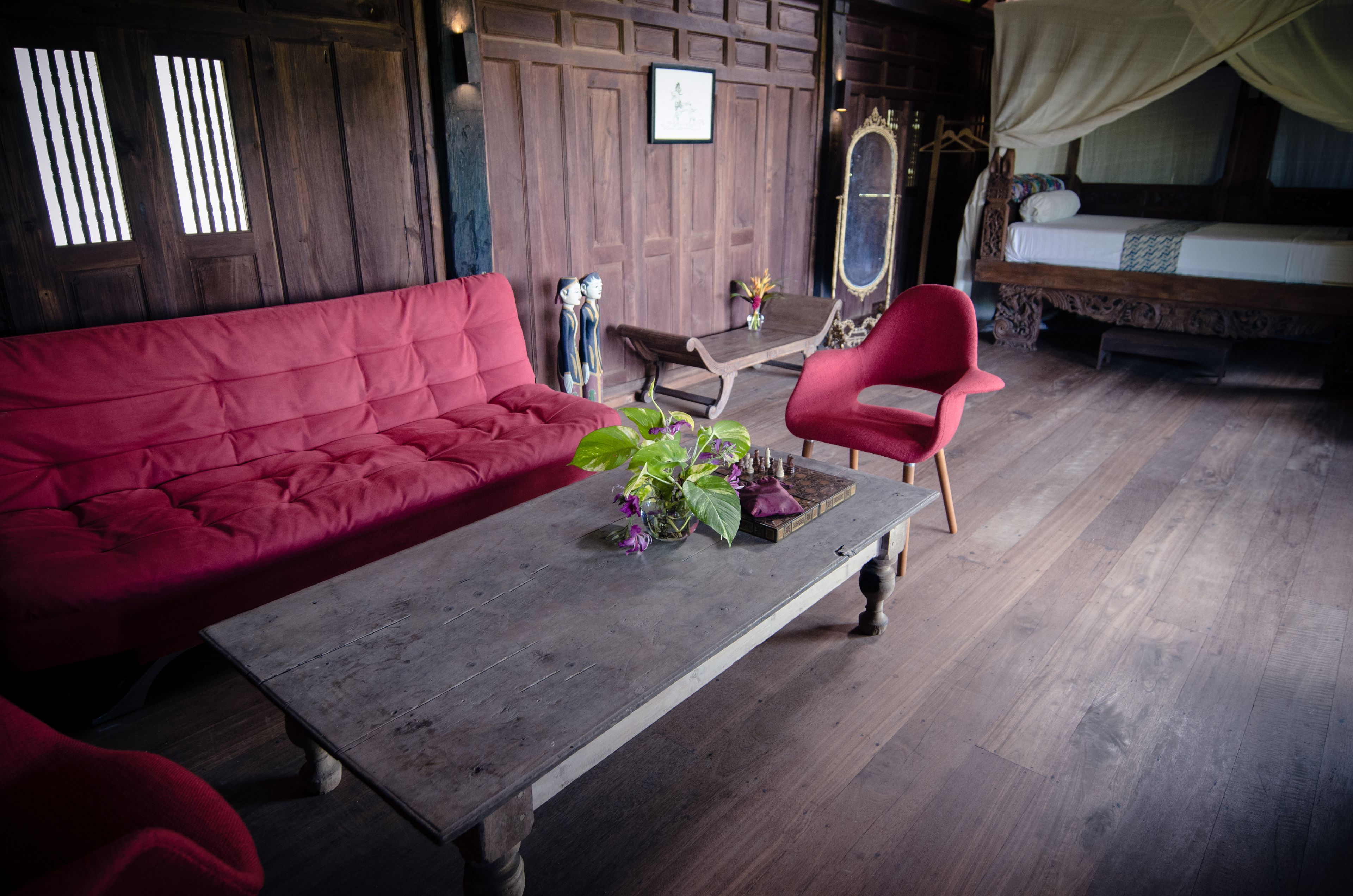 A room with a red sofa, a wooden coffee table with a vase of flowers, a red chair, and a bed with a canopy.