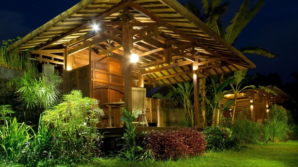 A wooden structure with a thatched roof, surrounded by lush greenery and illuminated by warm lighting. - Yabbiekayu Eco Bungalows (Sewon)