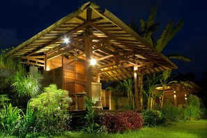 Una estructura de madera con techo de paja, rodeada de exuberante vegetación e iluminada con luz cálida.