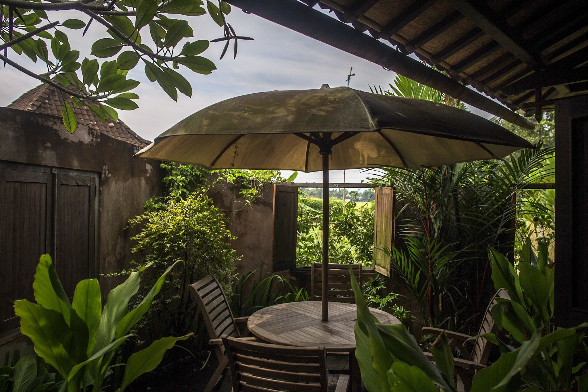 An outdoor patio with a wooden table and chairs, surrounded by lush greenery and a large umbrella providing shade.