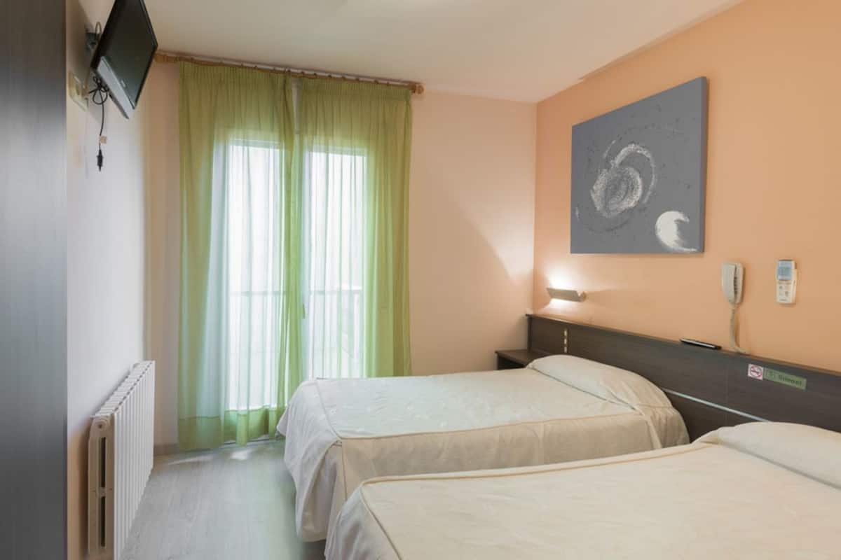 Habitació d'hotel amb dos llits, un televisor penjat a la paret, un quadre i una finestra amb cortines verdes.