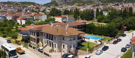 A stone building with a swimming pool, surrounded by parked cars and a residential area with red-roofed houses.