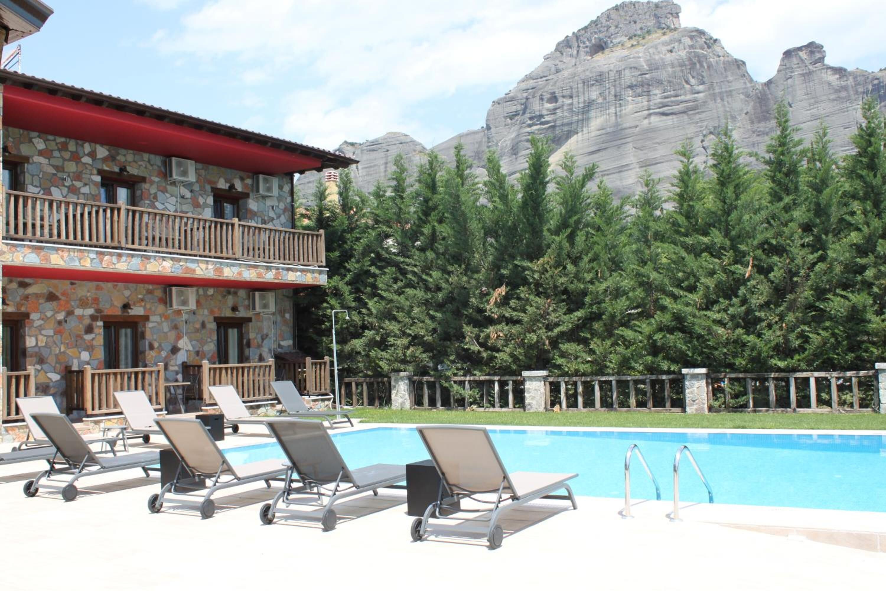 Un área de piscina con sillones de descanso, un edificio de piedra con tejado rojo y montañas al fondo.