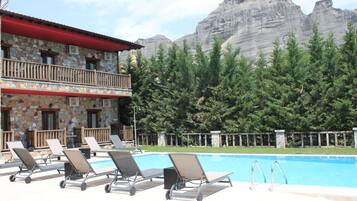 Un área de piscina con sillones de descanso, un edificio de piedra con tejado rojo y montañas al fondo.