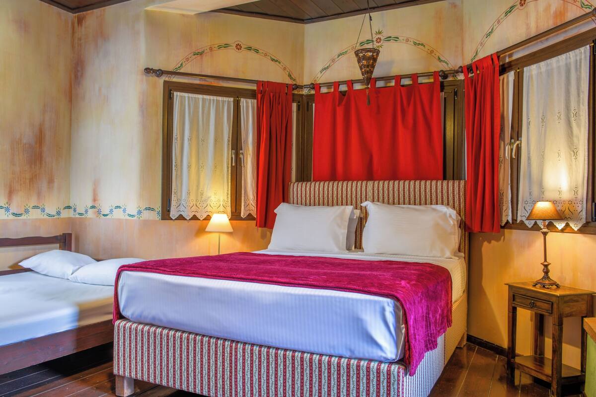 A hotel room with two beds, a red curtain, a patterned bedspread, and a small table with a lamp.