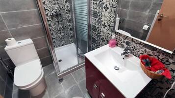 A bathroom with a toilet, a shower with patterned tiles, a sink with a mirror, and a red towel basket.