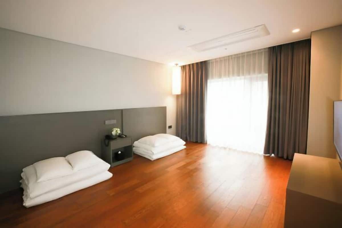 Ein Hotelzimmer mit Holzboden, einem Bett mit weißen Leinentüchern, einem Nachttisch, einer Lampe und einem großen Fenster mit Vorhängen.