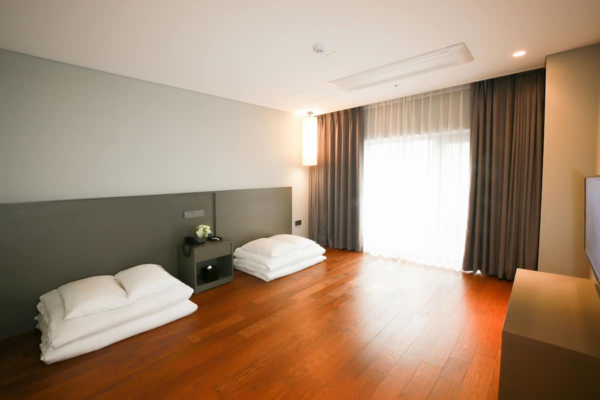 Ein Hotelzimmer mit Holzboden, einem Bett mit weißen Leinentüchern, einem Nachttisch, einer Lampe und einem großen Fenster mit Vorhängen.