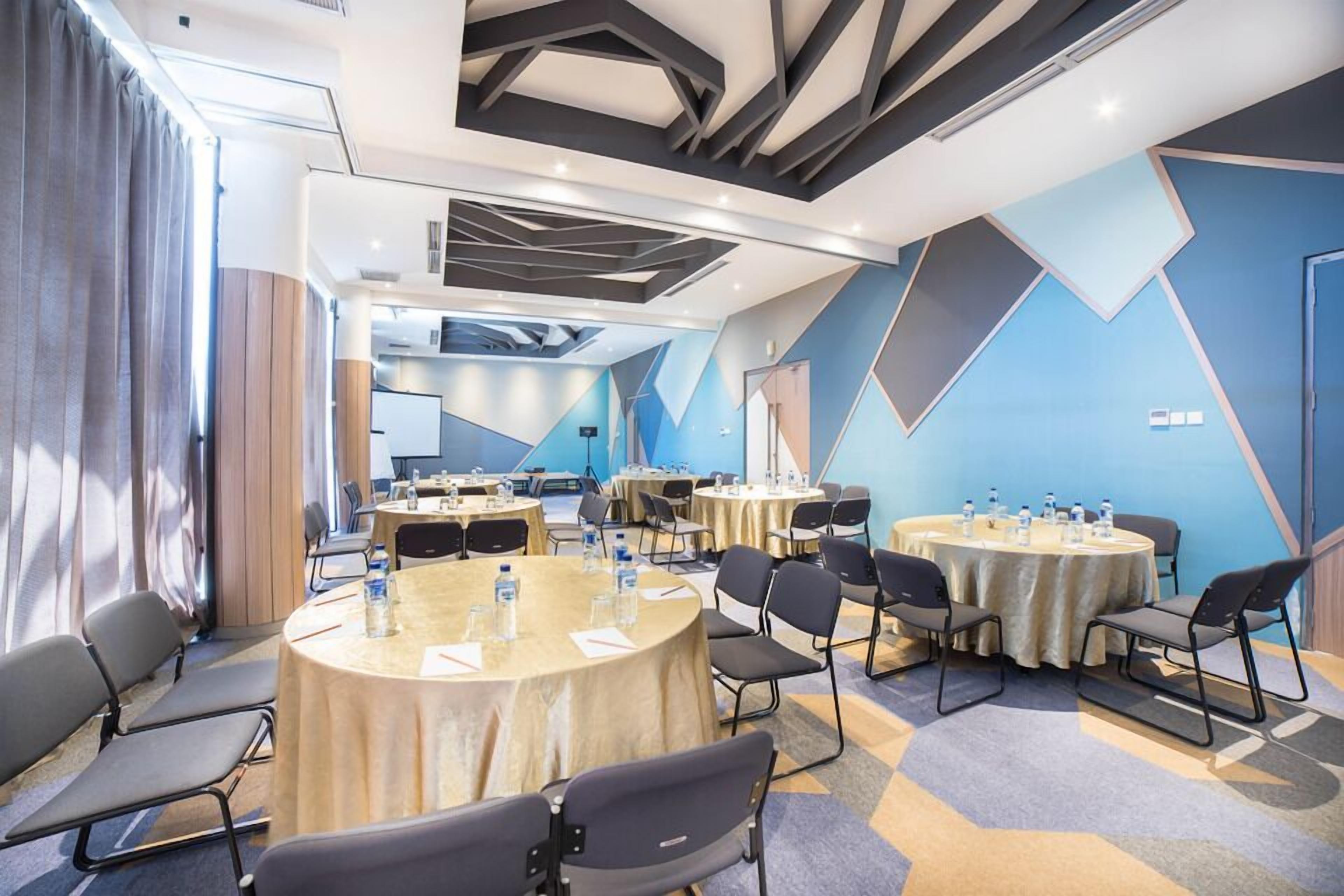 Un salón de conferencias con varias mesas redondas, cada una con sillas y botellas de agua. La sala presenta diseños geométricos en las paredes y un techo moderno.