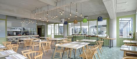 A spacious dining area with multiple tables and chairs, a kitchen in the background, and hanging pendant lights.