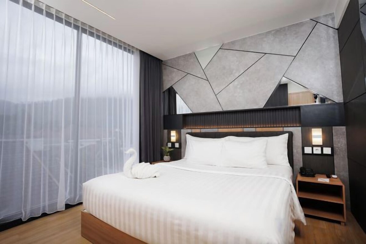 A modern hotel room with a large bed, a bedside table, and a view of the mountains through sheer curtains.