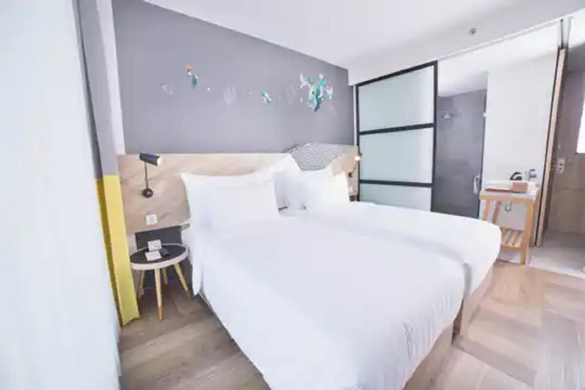 A modern hotel room with a large bed, a bedside table, a small round table, and a wardrobe.