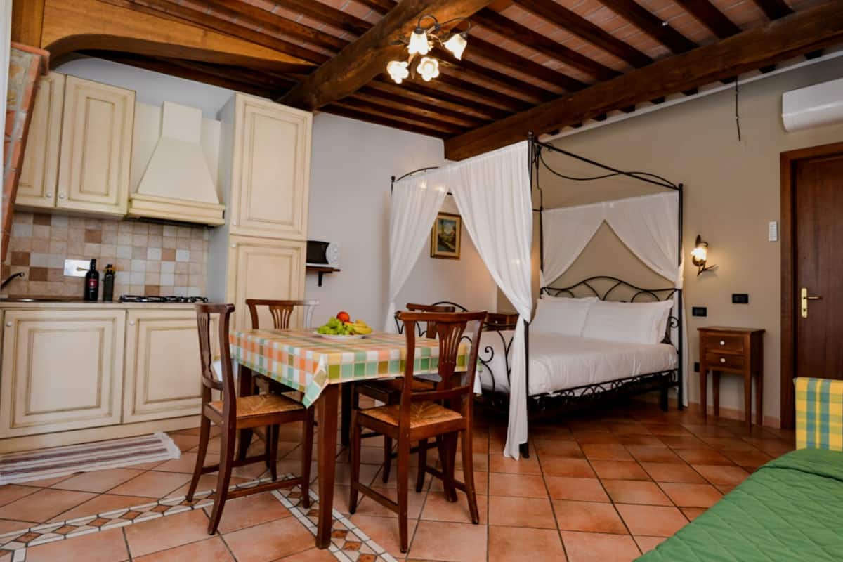 A bedroom with a four-poster bed, a dining table with chairs, and a kitchen area with cabinets and a stove.