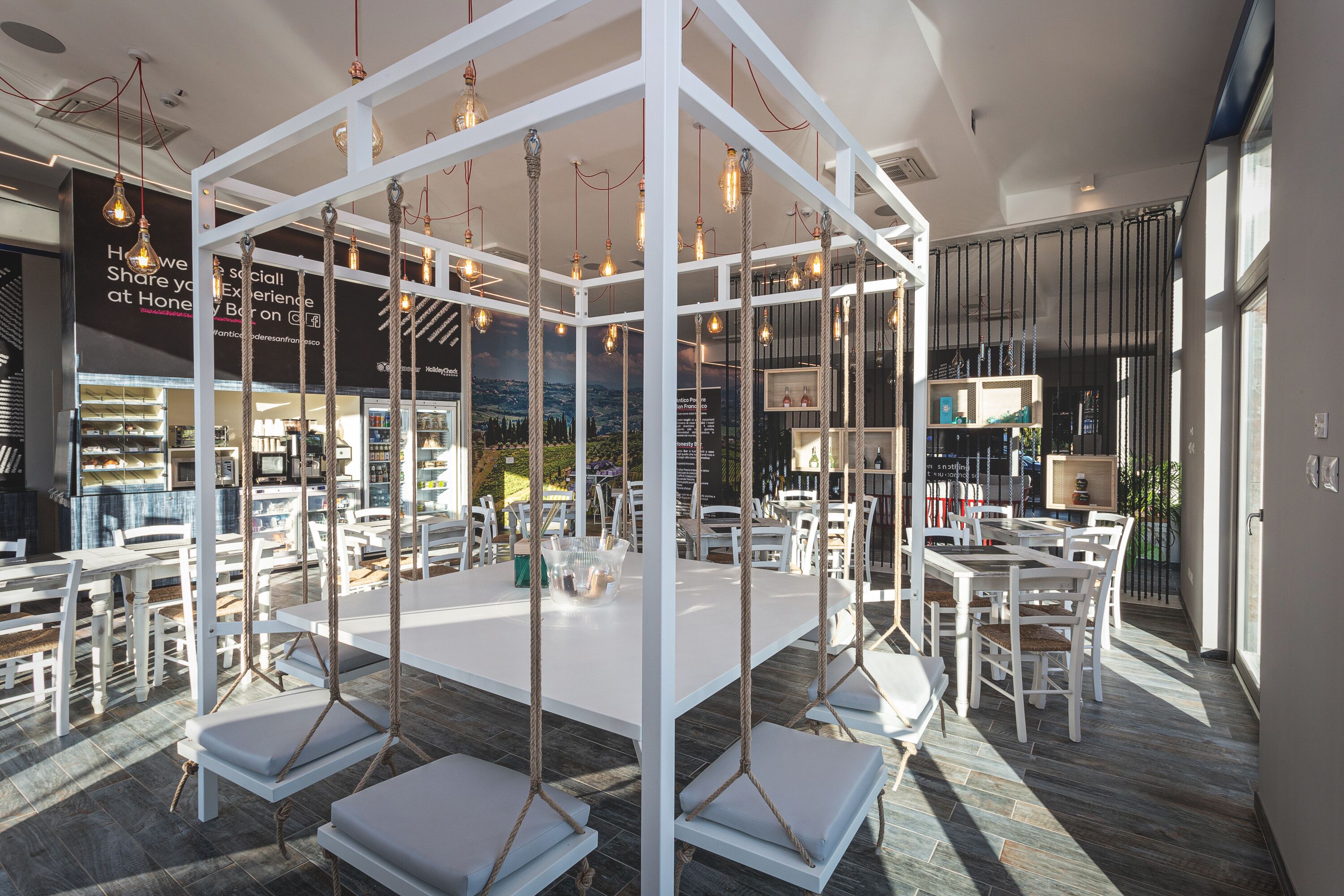Un espace repas moderne, avec des meubles blancs et des balançoires en corde. Les tables sont disposées autour d’une structure centrale blanche, et des lampes sont suspendues au plafond.