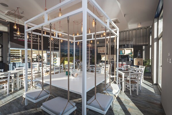 Comedor moderno con mobiliario blanco y columpios de cuerda. Las mesas están dispuestas alrededor de una estructura blanca central, y hay luces colgantes.