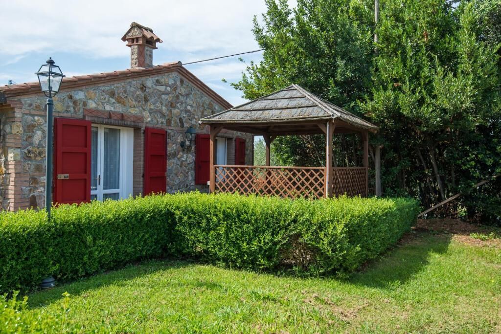 A stone building with red shutters, a wooden gazebo, and a well-maintained hedge.