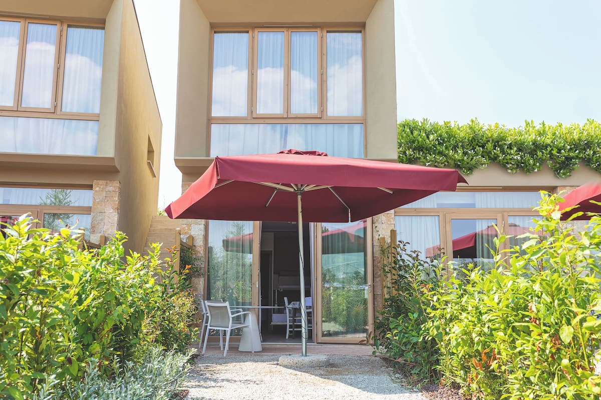 An outdoor patio with a table and chairs under a red umbrella, surrounded by greenery and a building with large windows.
