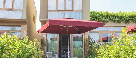 An outdoor patio with a table and chairs under a red umbrella, surrounded by greenery and a building with large windows.