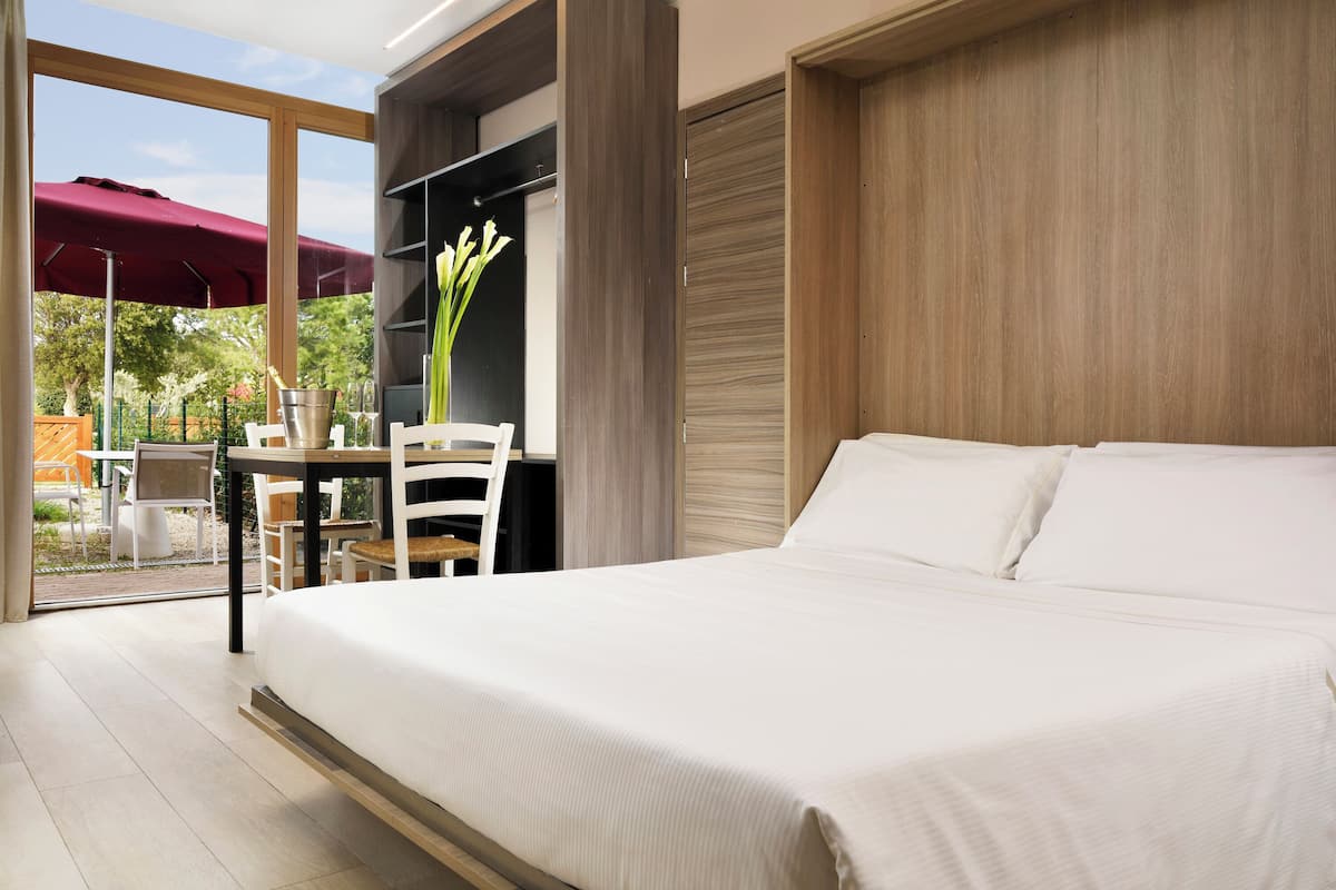 A modern bedroom with a large bed, a small table with chairs, a potted plant, and a sliding door leading to an outdoor area.