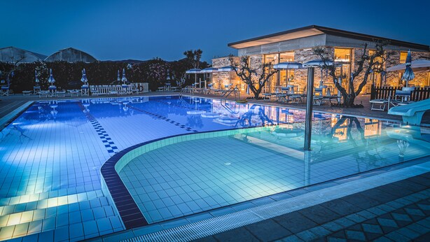 A swimming pool with lounge chairs and umbrellas, surrounded by a stone wall and a building with large windows. - San Francesco Country Resort (Rosignano Marittimo)