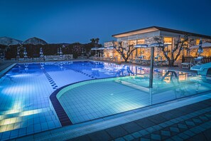 Una piscina con sillas de descanso y sombrillas, rodeada por un muro de piedra y un edificio con amplias ventanas.