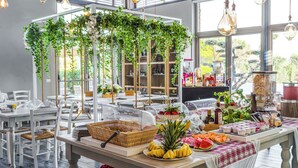 A buffet setup with various food items, including fruits and bread, in a well-lit room with large windows and hanging light fixtures. - San Francesco Country Resort (Rosignano Marittimo)