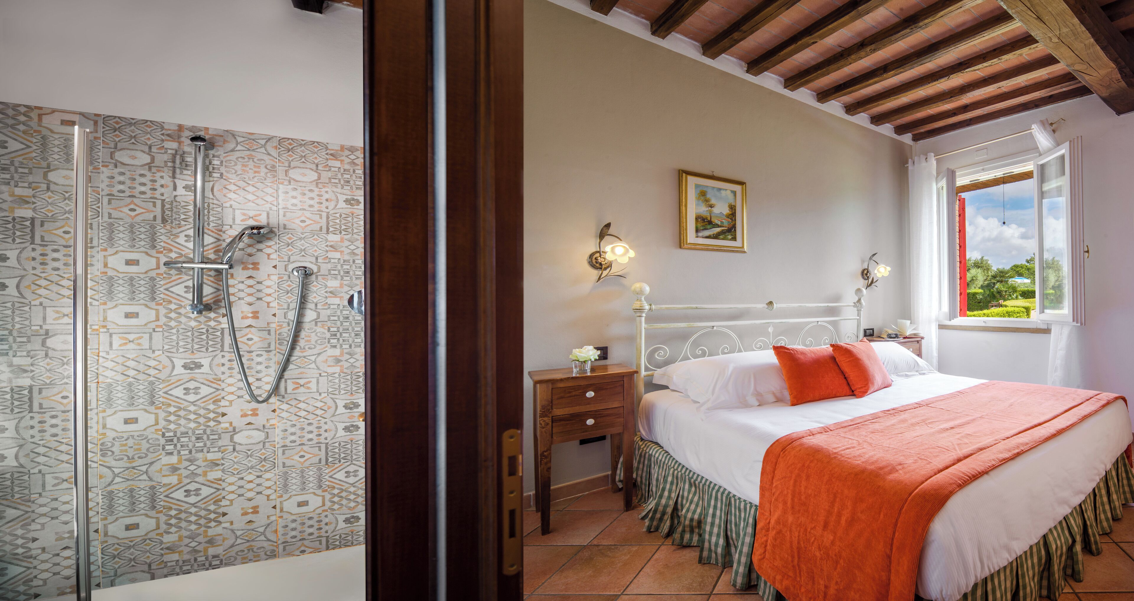 A bedroom with a bed, a nightstand, a bedside table, a window with a view, and a shower area with patterned tiles.