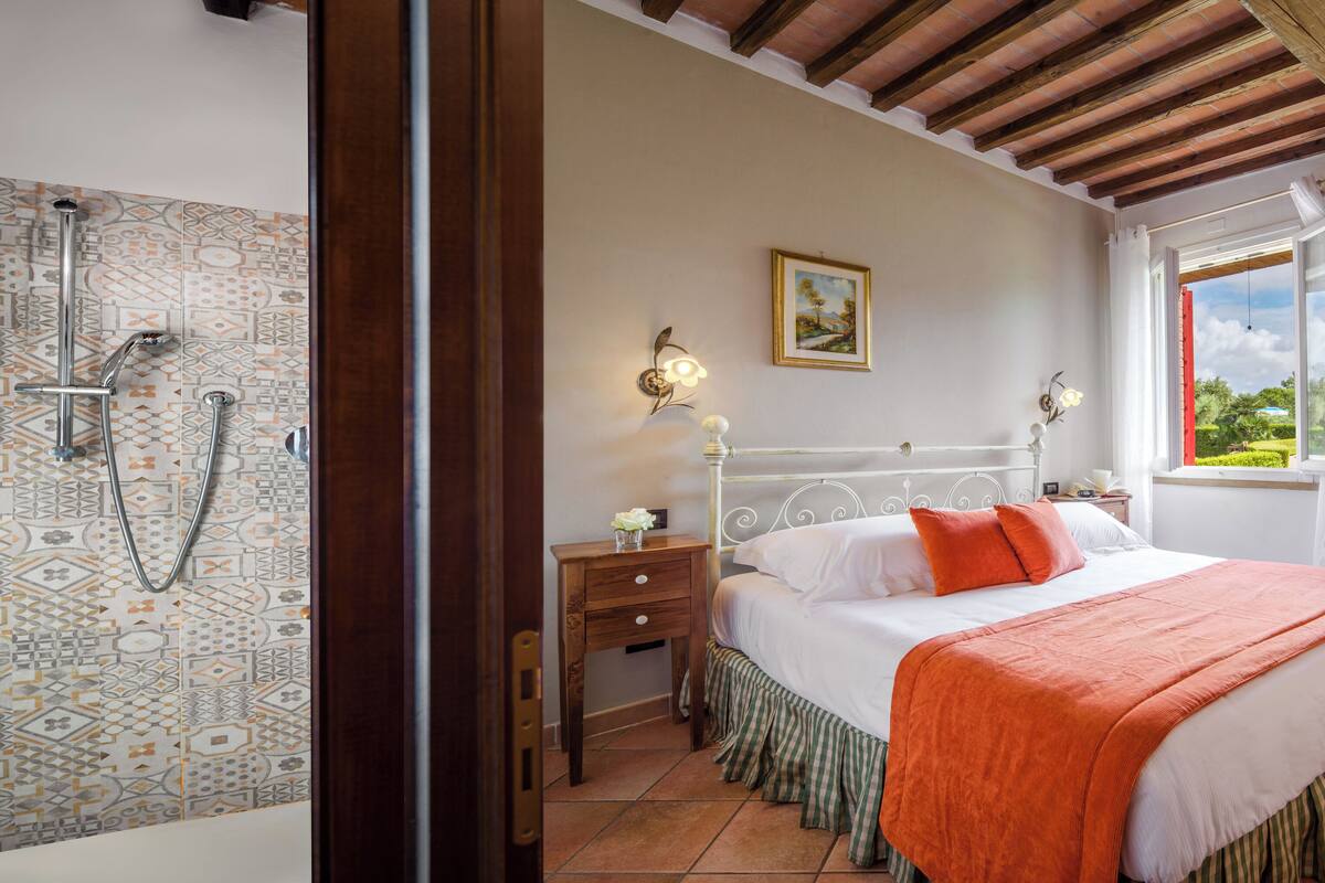 A bedroom with a bed, a nightstand, a bedside table, a window with a view, and a shower area with patterned tiles.