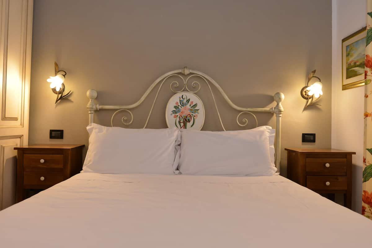 A bed with a decorative headboard, two bedside tables with lamps, and a floral painting on the wall.
