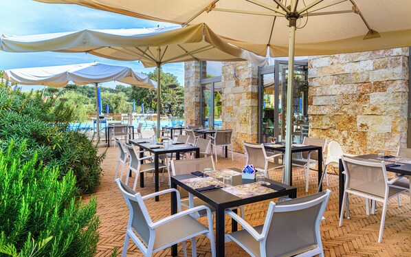 Un área de comedor al aire libre con mesas y sillas, una pared de piedra y una piscina al fondo.