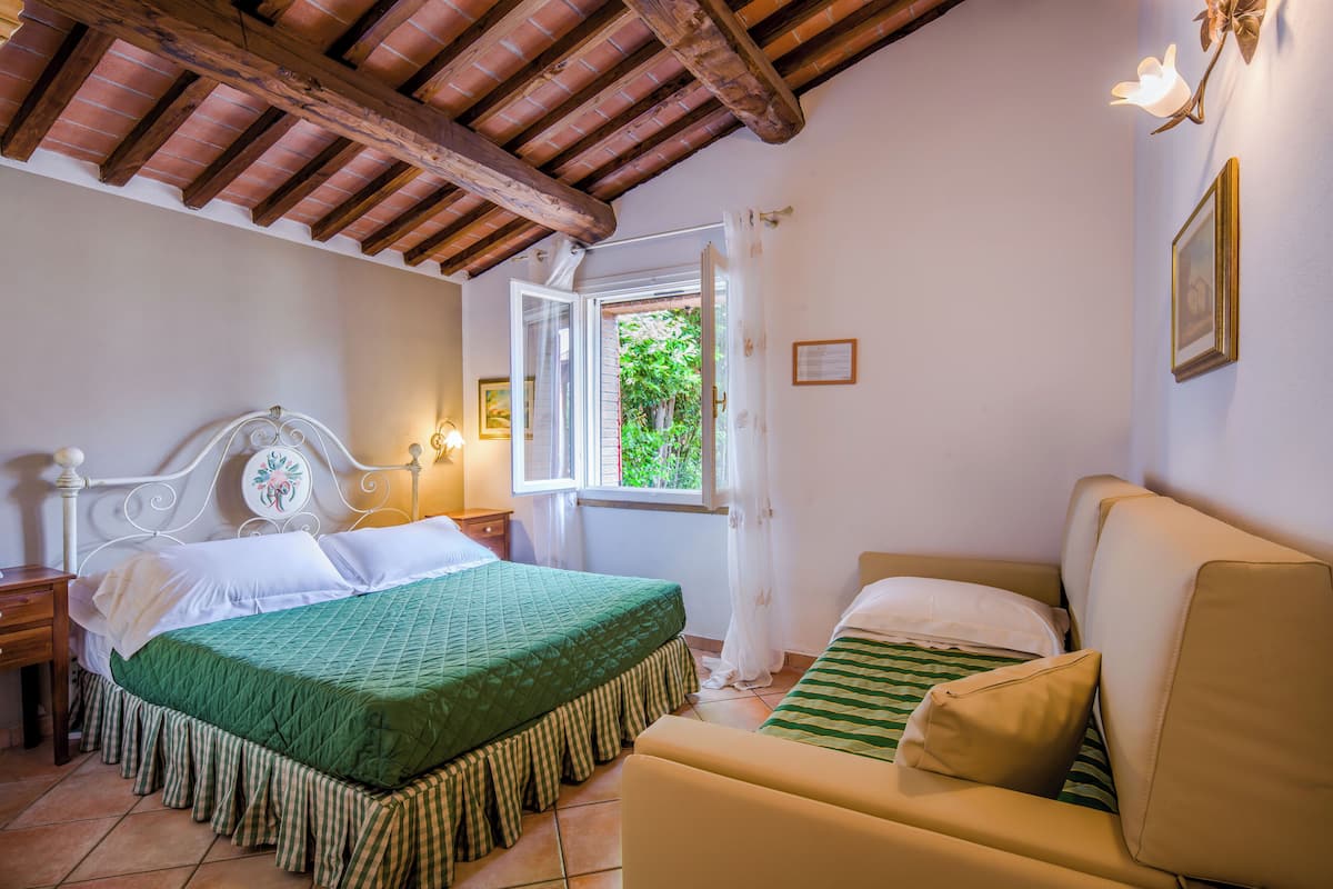 A bedroom with a bed, a sofa, a nightstand, a window with curtains, and a wooden ceiling.