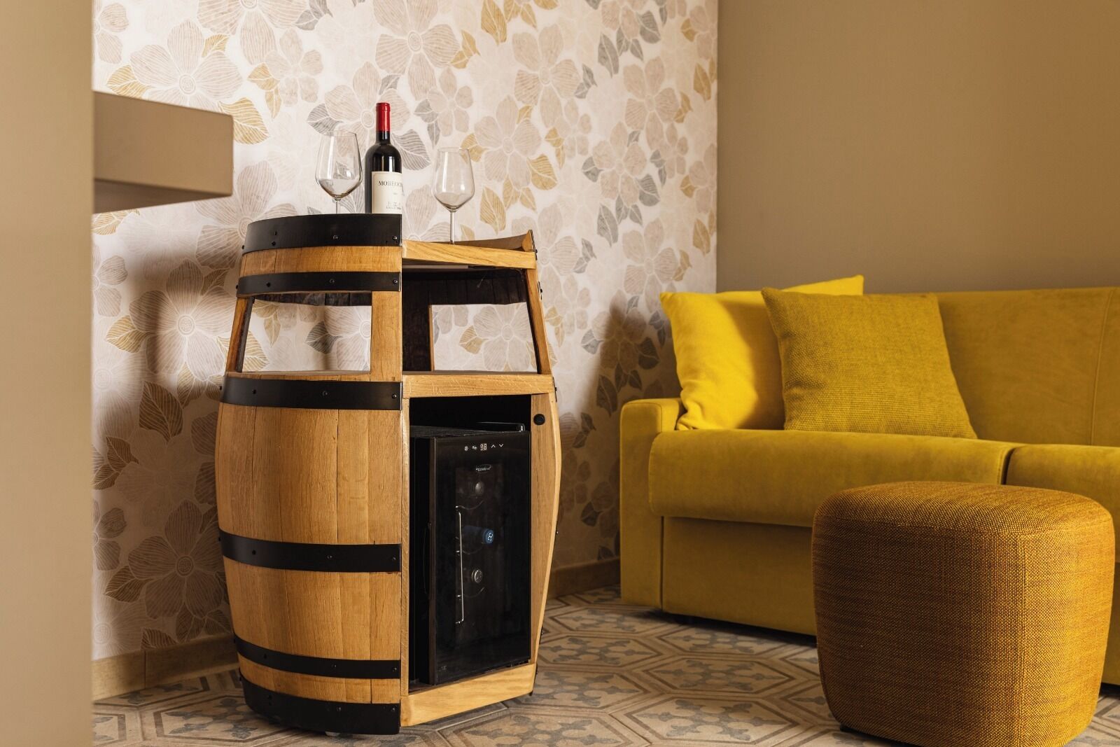 A wooden wine barrel-shaped cabinet with a wine bottle and glasses on top, a modern black dishwasher, a yellow sofa, and a woven ottoman.