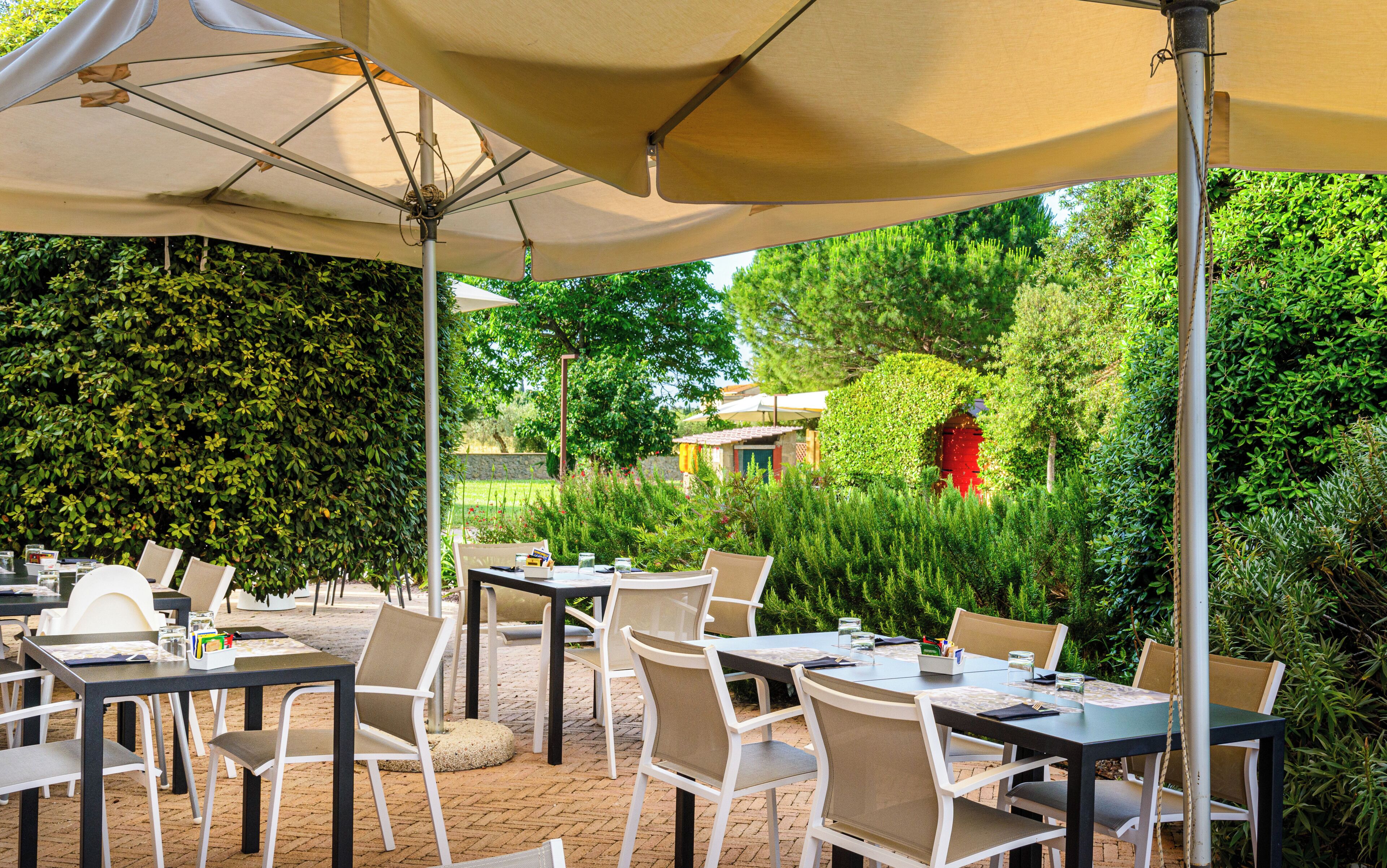 Un espace de restauration en plein air, avec des tables et des chaises placées sous des parasols, entouré d’une végétation luxuriante et d’un mur en pierre.