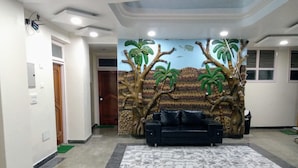 A lobby with a black leather sofa, a marble-patterned rug, and a wall mural of trees and a landscape. - Hotel Raghunath (Jammu)