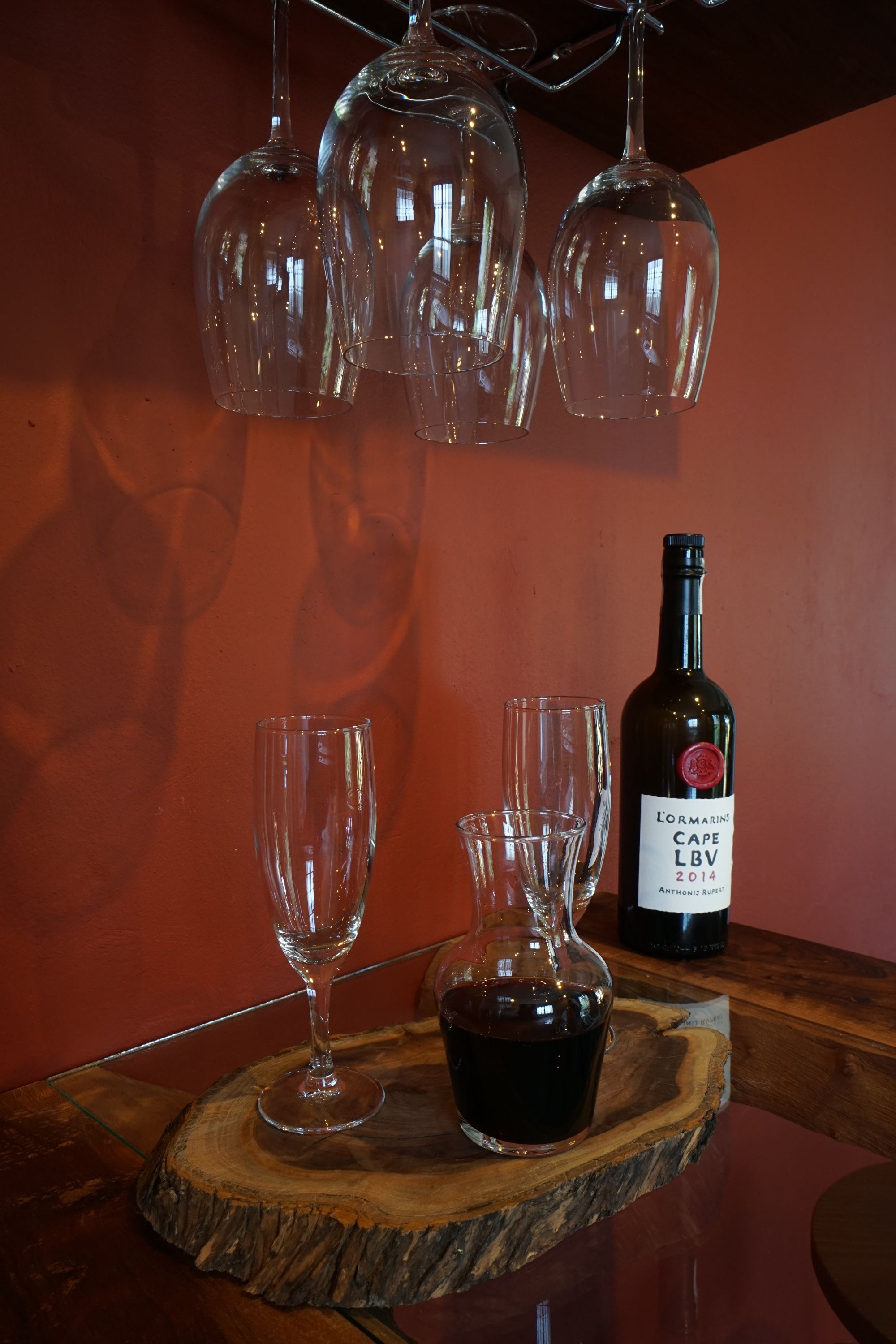 A wine bottle on a wooden coaster with two glasses, one filled with wine, and several empty glasses hanging above.