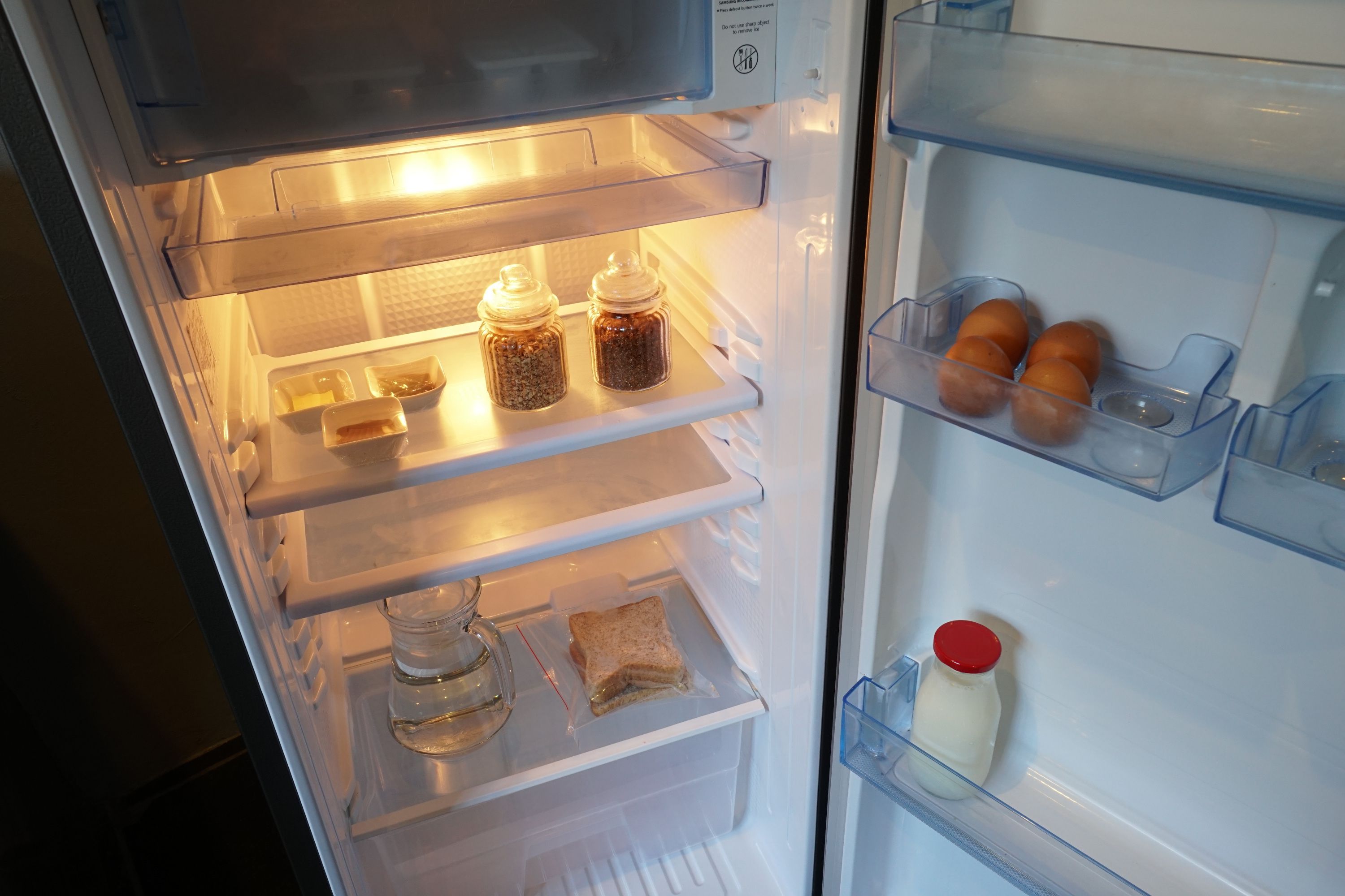 Un refrigerador abierto con repisas que contienen frascos, un cartón de leche y un sándwich.