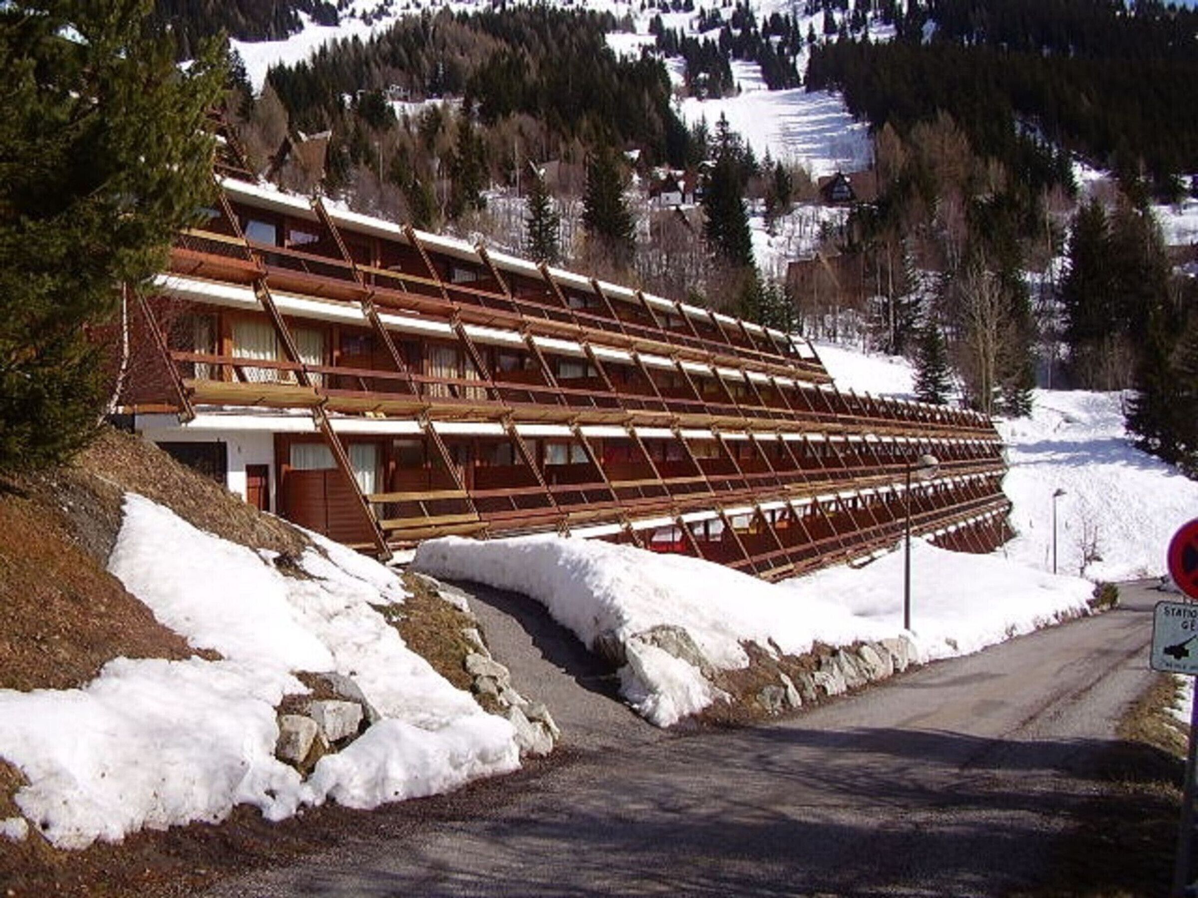A large wooden building with a sloped roof, situated on a snowy hillside with a road in front.