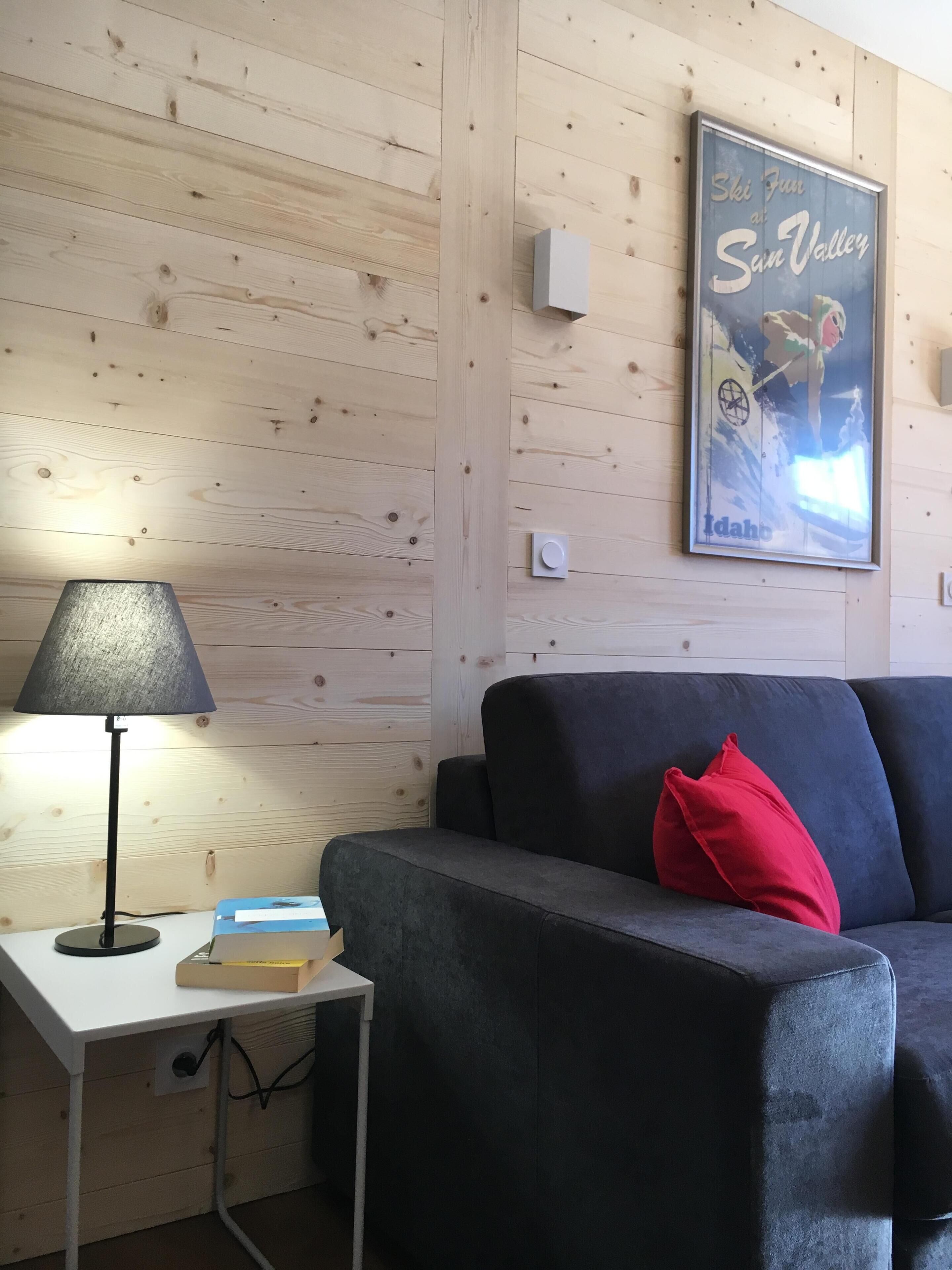 Ein Zimmer mit einer holzgetäfelten Wand, einem dunkelgrauen Sofa mit einem roten Kissen, einem kleinen weißen Tisch mit einer Lampe und Büchern sowie einem gerahmten Poster an der Wand.