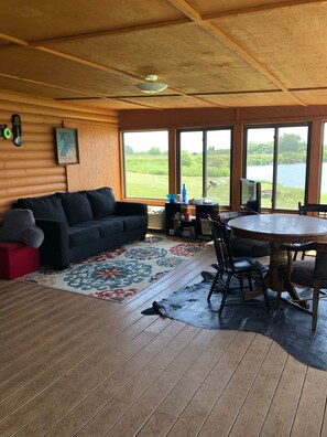 A cozy living room with a dark blue sofa, a wooden dining table with chairs, and a large window offering a view of a lake and greenery. - Cabin on Private Lake (Allen)