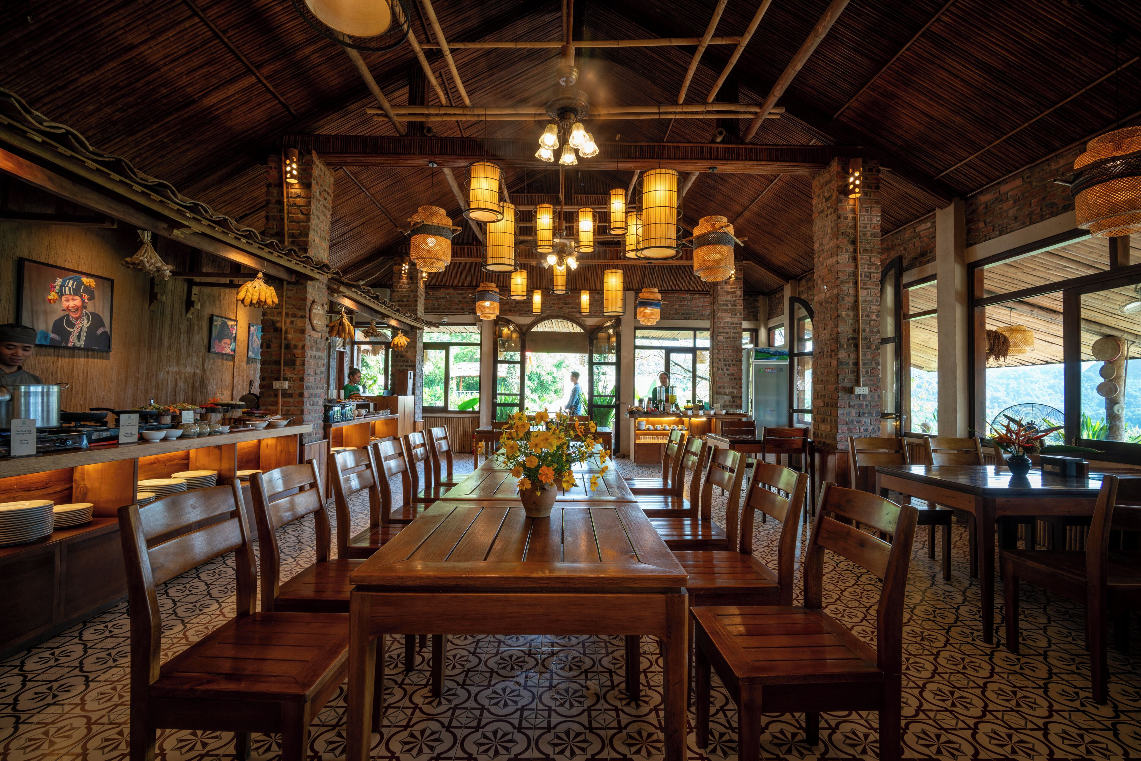 A restaurant with a long wooden table, multiple chairs, hanging lights, and a brick wall.