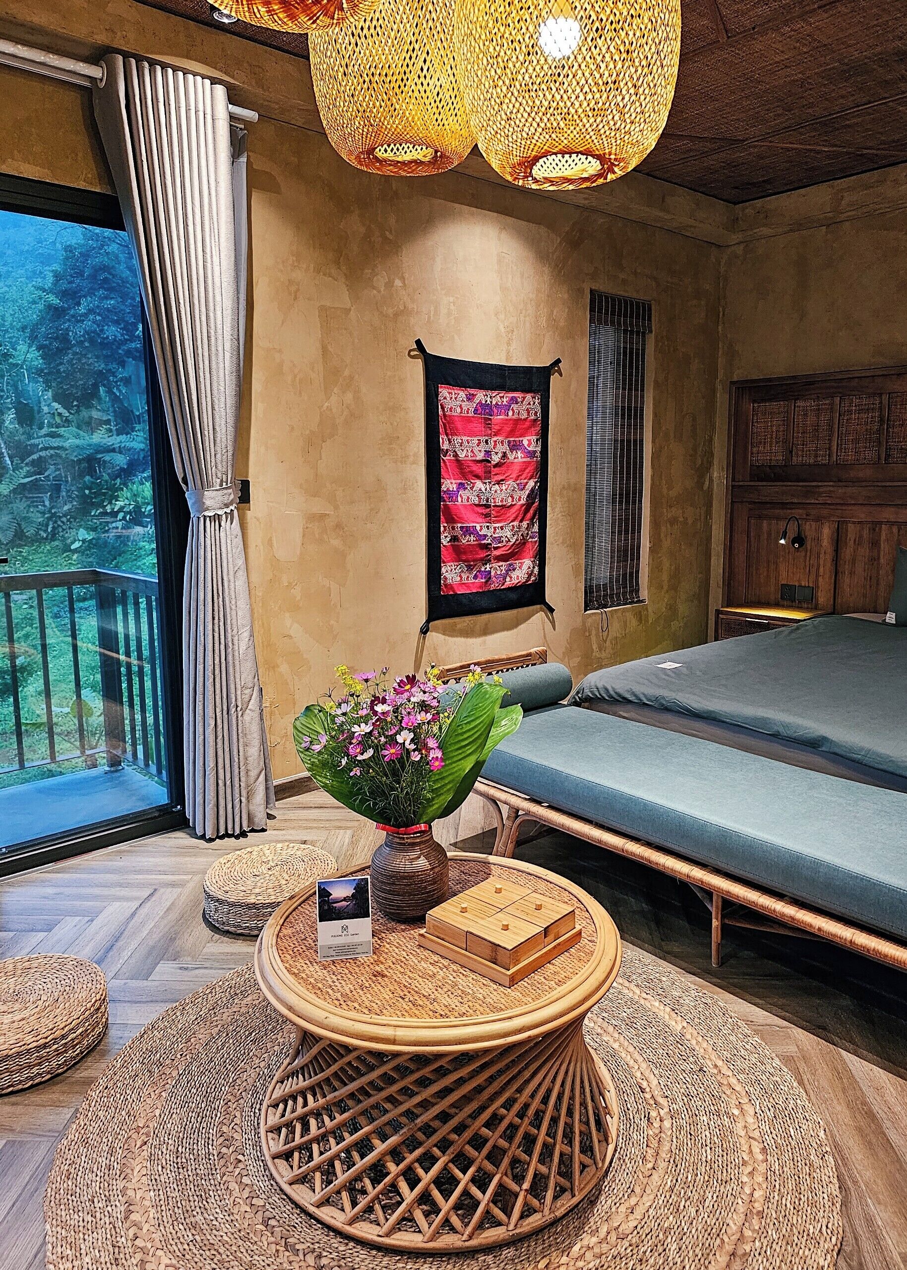 Um quarto com uma cama, uma janela com cortinas, uma tapeçaria na parede, uma mesa de vime com um vaso de flores e uma cabeceira de madeira.