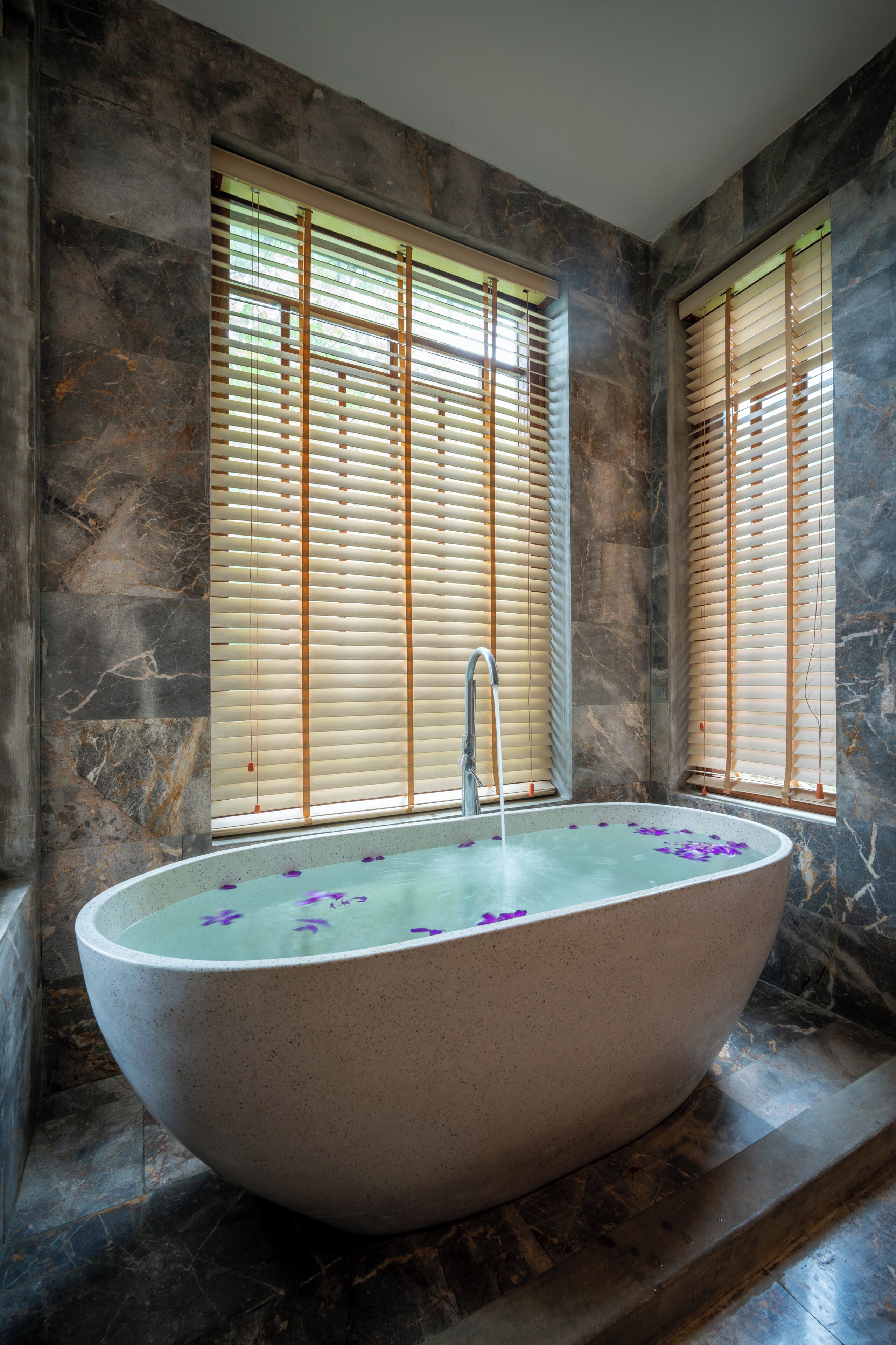 A large freestanding bathtub with water and flower petals, set against a wall with a stone-like texture.