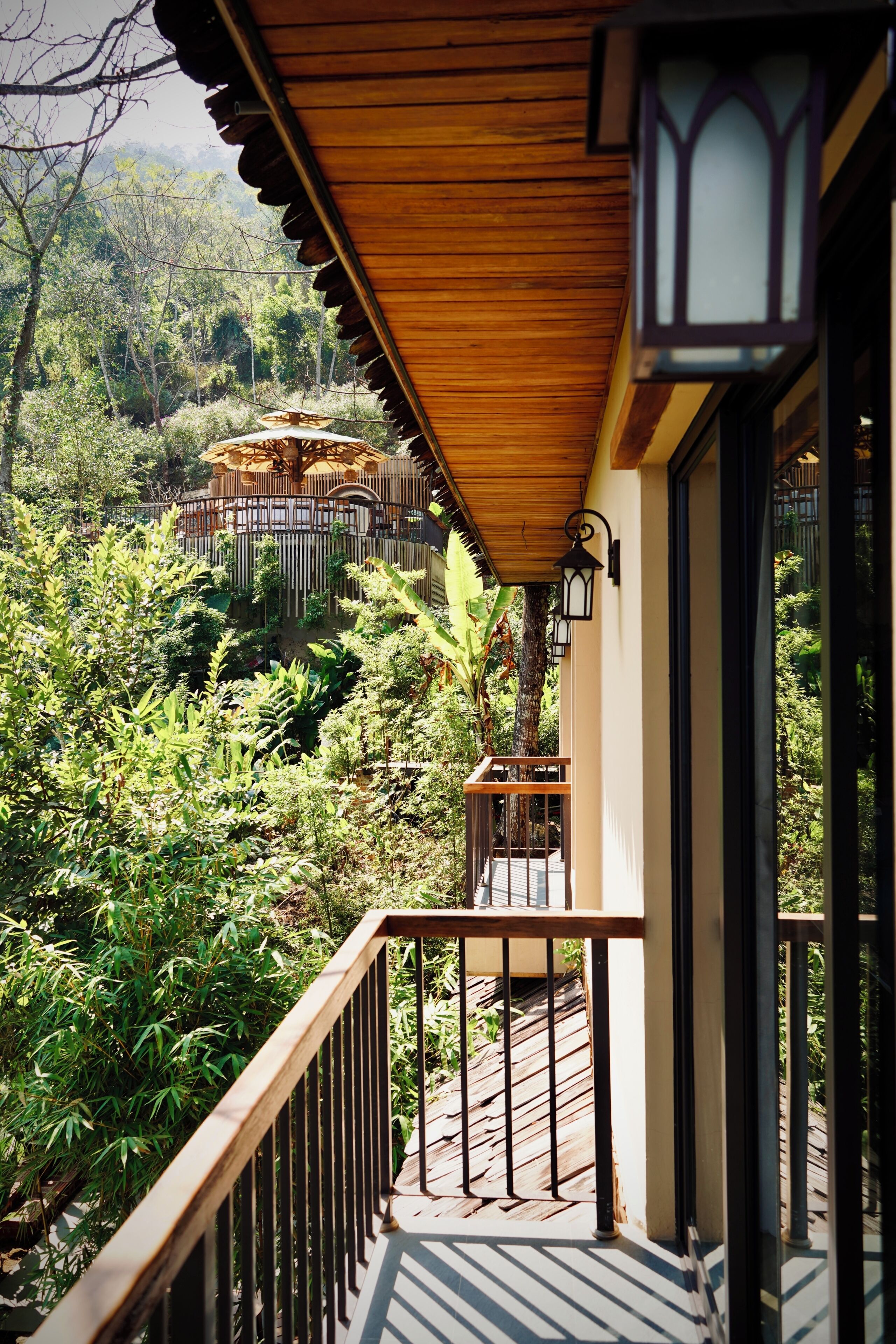 A wooden balcony with a railing overlooking a lush garden and a traditional building in the background.