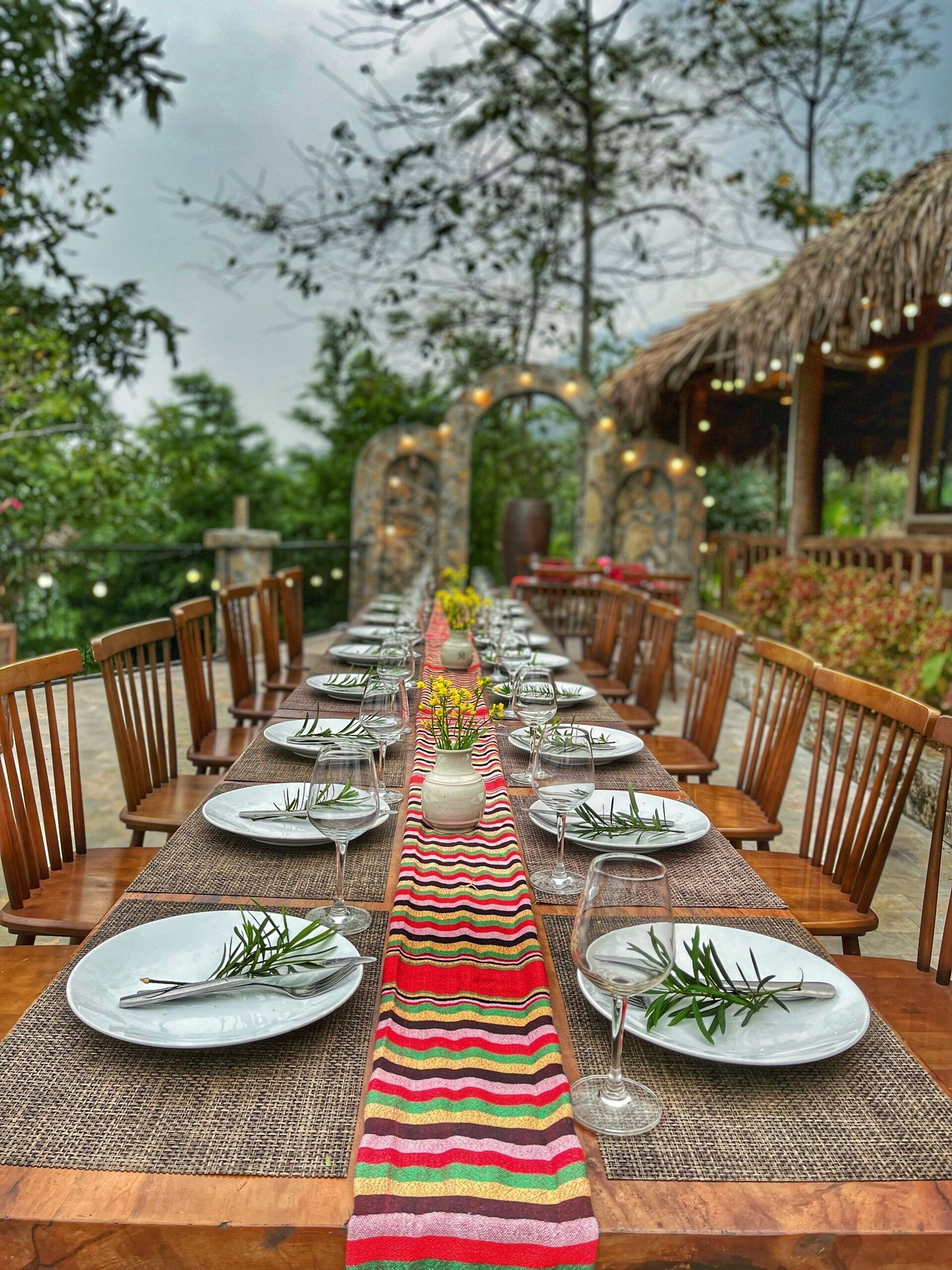 A long dining table set for a meal with chairs on both sides, a decorative centerpiece, and a thatched roof structure in the background.