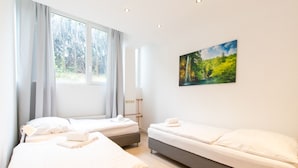 A hotel room with two beds, a large window with curtains, a painting on the wall, and a bedside table with a lamp. - Representative City Villa (Meerbusch)