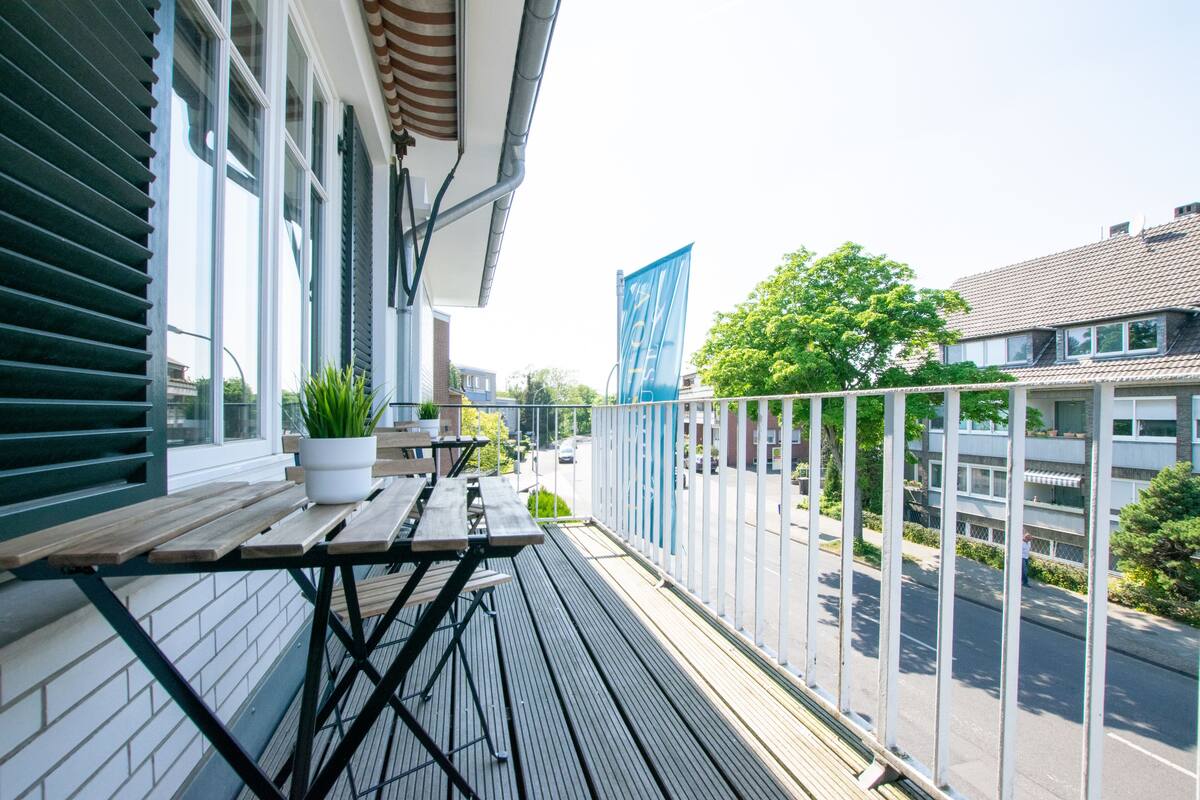 Un balcon avec une table et des chaises en bois, des plantes en pot et une vue sur la rue et les immeubles.