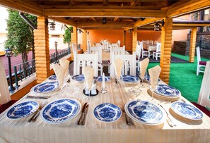 A covered outdoor dining area with tables set for a meal, featuring blue and white patterned plates and white chairs.